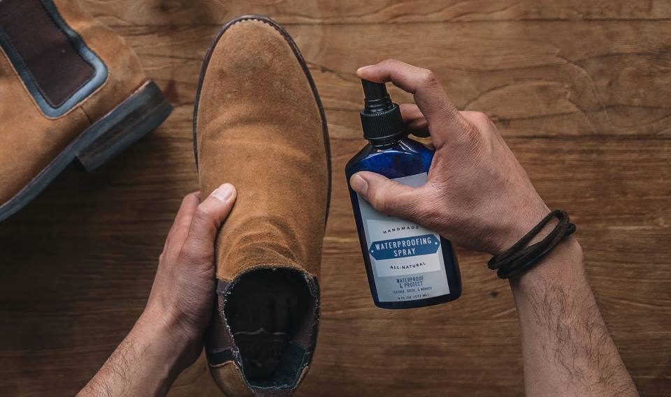 A close-up of a hand using a soft cloth to rub mink oil onto tan suede boots, protecting them from water damage
