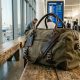 A durable canvas travel duffel bag rests on a bench inside an airport.